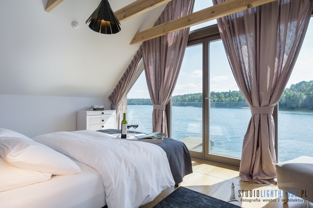 Jasna sypialnia na poddaszu z dużym oknem na jezioro, butelka wina i kieliszki na łóżku, drewniane belki stropowe, nowoczesna lampa wisząca.