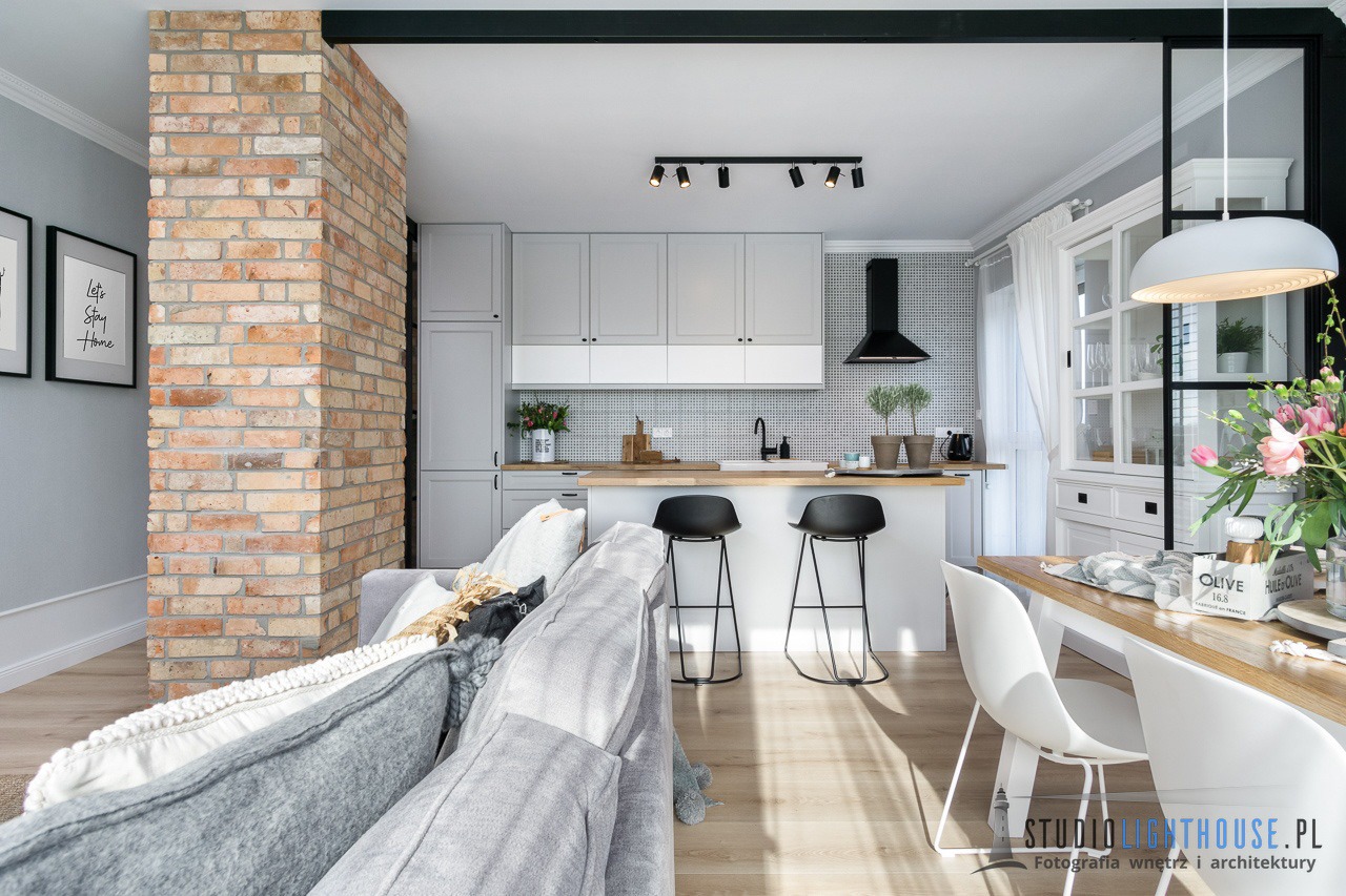 Jasne, przestronne wnętrze z ceglanym filarem, szarą kuchnią z wyspą i czarnymi hokerami, oraz częścią wypoczynkową z szarą sofą. Na pierwszym planie stół z białymi krzesłami i bukietem kwiatów.