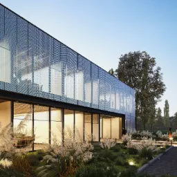 Nowoczesny budynek biurowy z perforowaną elewacją i bujną roślinnością ozdobną, oświetloną punktowymi lampami, widziany z perspektywy niskiego kąta.