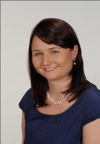 Portret uśmiechniętej kobiety z brązowymi włosami, w granatowej bluzce i perłowym naszyjniku, na jasnym tle.