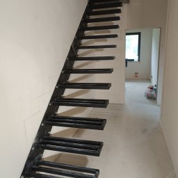 SŁAWKO-STAL - Surowe, czarne schody metalowe z prostokątnymi stopniami, zamontowane przy białej ścianie w surowym wnętrzu. Widoczne ślady po montażu i okno w tle.