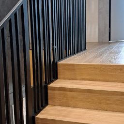 SŁAWKO-STAL - Nowoczesne schody z drewnianymi stopniami i czarną, minimalistyczną balustradą. Widok z dołu, podkreślający solidność wykonania i estetykę wykończenia.