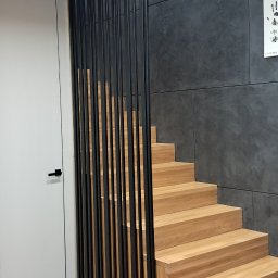 SŁAWKO-STAL - Drewniane schody z czarną, pionową balustradą w nowoczesnym wnętrzu. Jasna podłoga i ciemnoszara ściana tworzą kontrast. Minimalistyczny design.