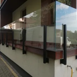 Nowoczesna szklana balustrada balkonowa z ciemnymi, metalowymi słupkami mocującymi do drewnianej podstawy, odbijająca otoczenie i błękitne niebo.