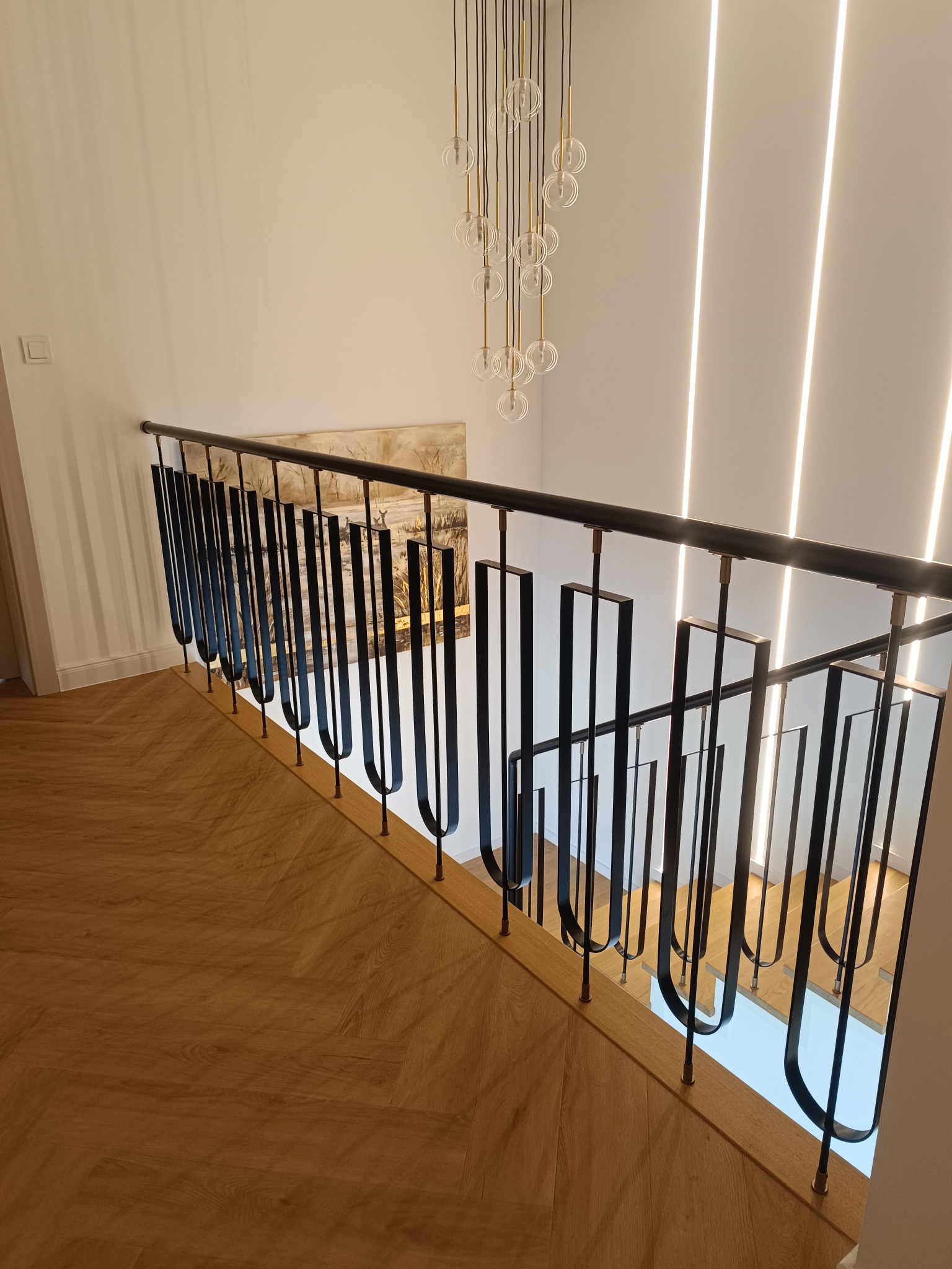 Nowoczesna balustrada metalowa z czarnymi, pionowymi elementami o zaokrąglonym kształcie, zamontowana na drewnianych schodach. W tle oświetlenie LED i obraz.