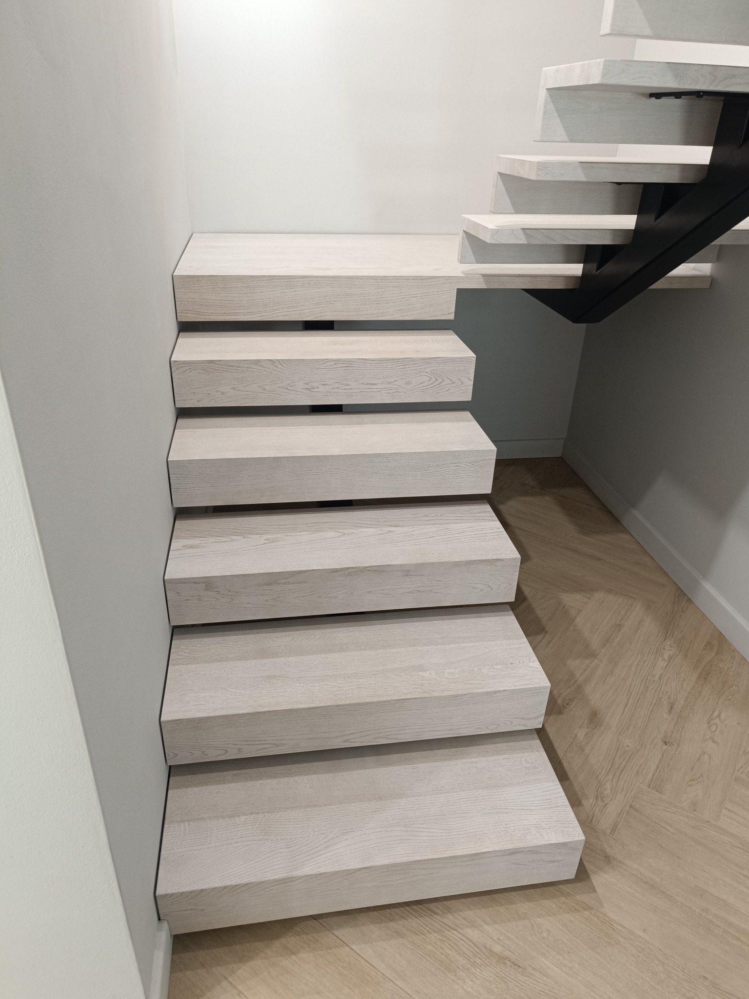 Minimalistyczne schody z drewnianymi stopniami w odcieniach bieli, wsparte na czarnej, metalowej konstrukcji. Parkiet w jodełkę na podłodze.