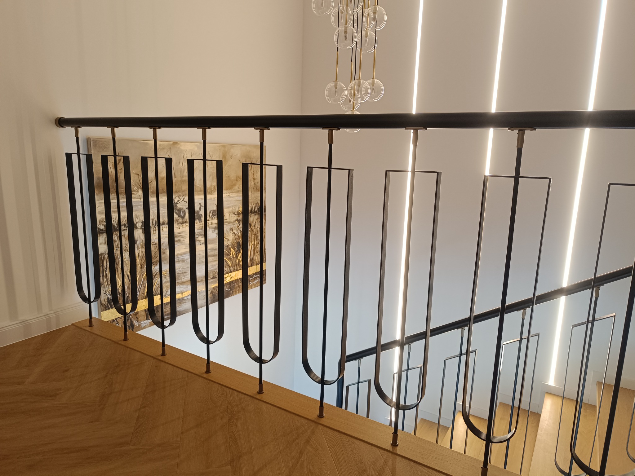 Nowoczesna balustrada schodowa z czarnymi, metalowymi elementami o unikalnym kształcie, na tle białych ścian i drewnianych schodów. Widoczny obraz na ścianie i designerska lampa.