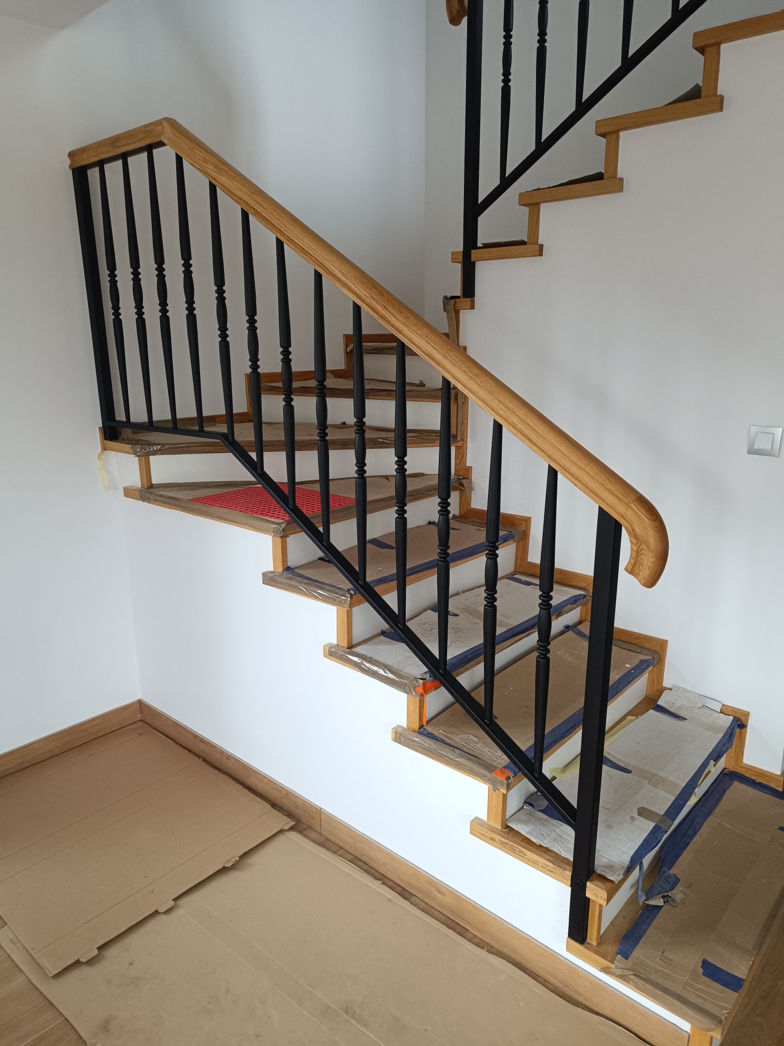Drewniane schody z czarną balustradą w trakcie montażu, zabezpieczone folią i kartonem. Widoczne drewniane stopnie i fragment ściany z włącznikiem.