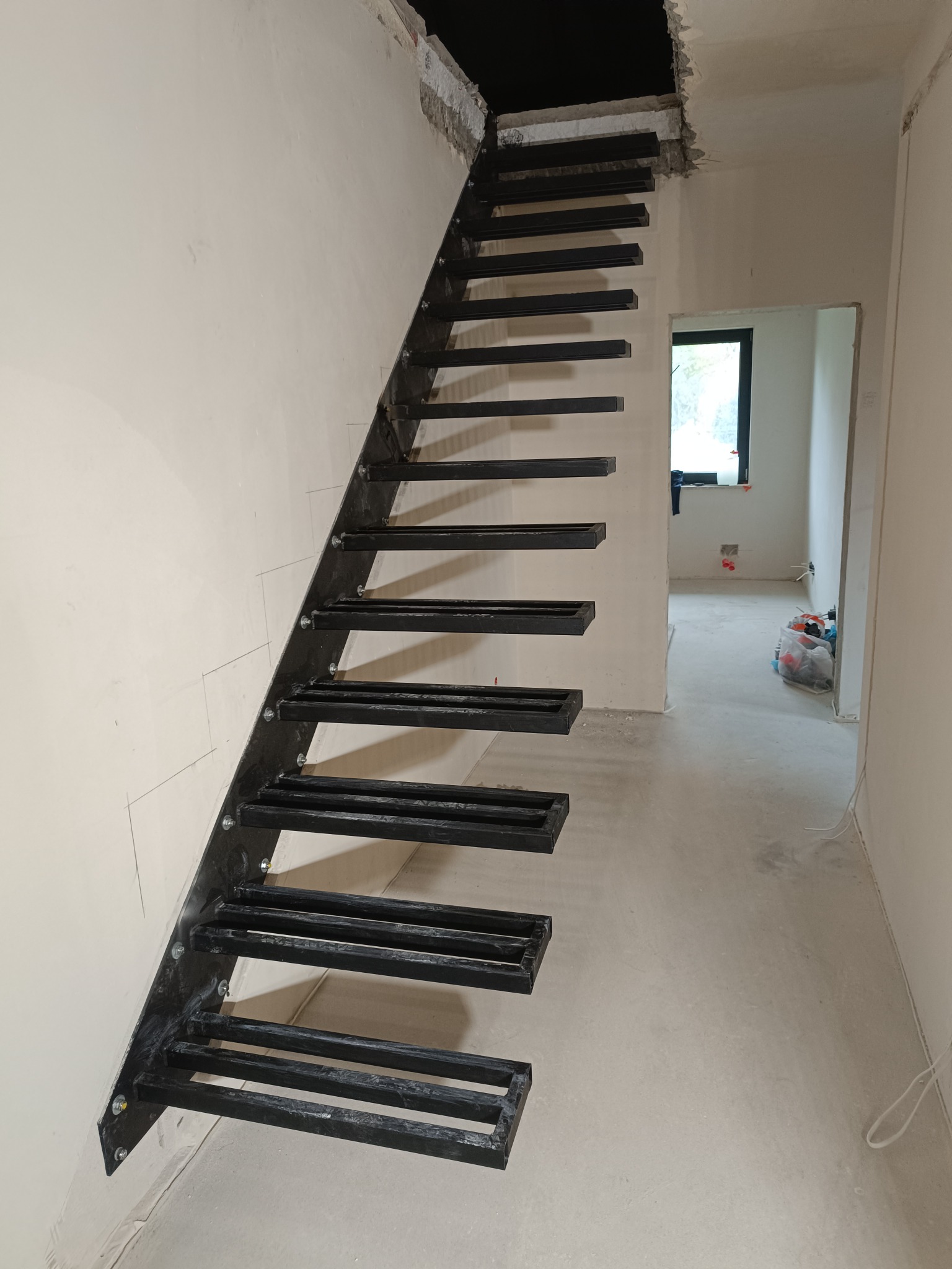 Surowe, czarne schody metalowe z prostokątnymi stopniami, zamontowane przy białej ścianie w surowym wnętrzu. Widoczne ślady po montażu i okno w tle.