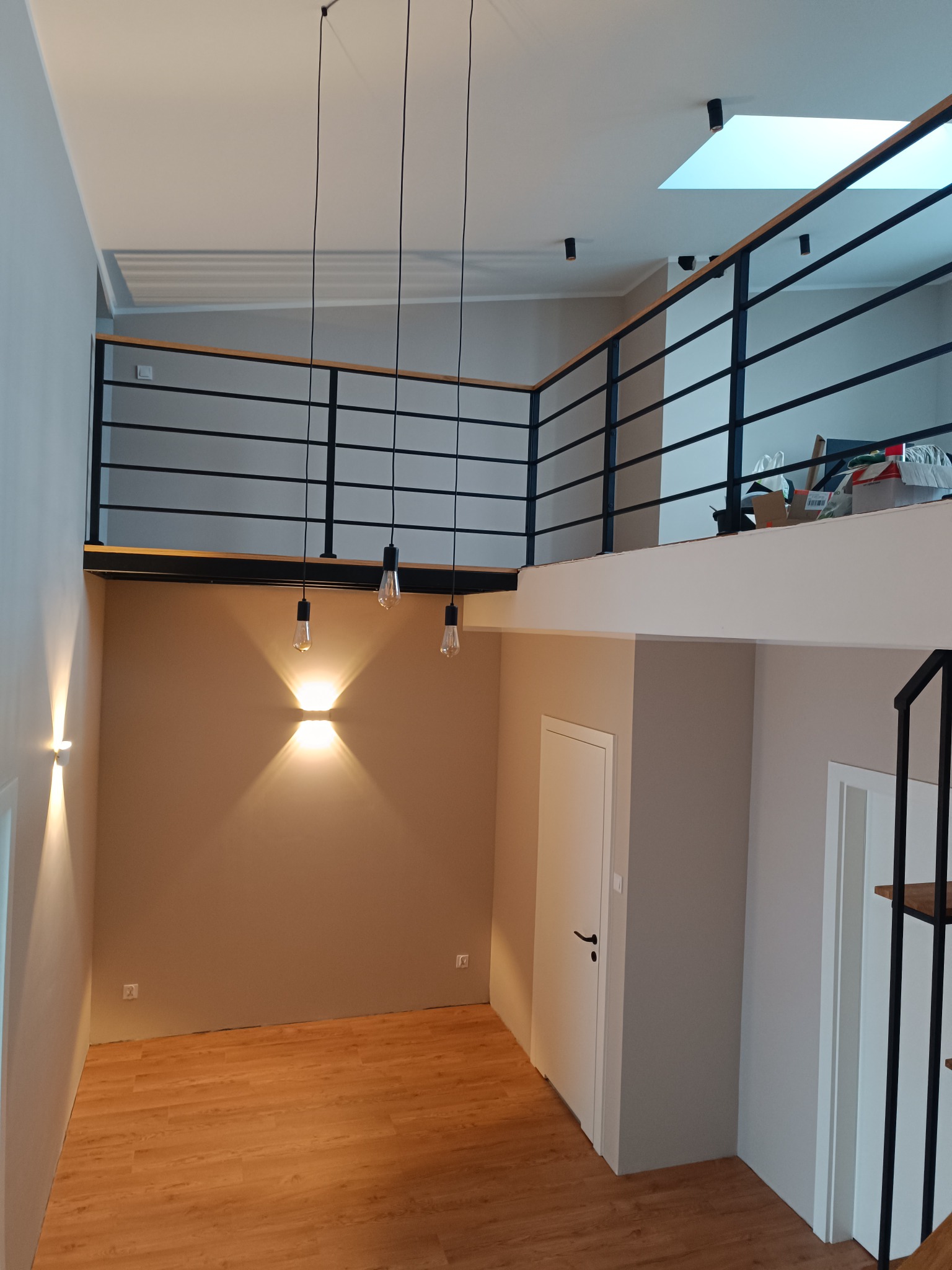 Dwupoziomowe wnętrze z czarną balustradą i drewnianymi elementami. Widoczne schody, oświetlenie punktowe i lampy wiszące. Minimalistyczny design z jasnymi ścianami i drewnianą podłogą.