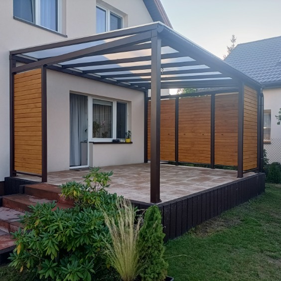 Zadaszona, drewniana pergola z metalową konstrukcją, przylegająca do domu. Widoczne drewniane panele boczne i kamienna podłoga tarasu. Ujęcie z poziomu ogrodu.