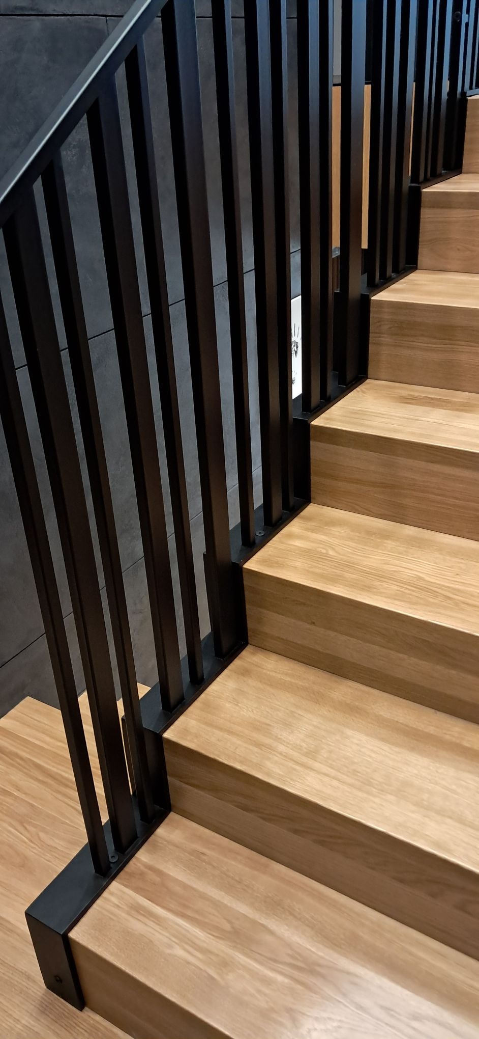 Nowoczesne schody z drewnianymi stopniami i czarną, metalową balustradą o minimalistycznym designie. Ujęcie z dołu, podkreślające geometrię i materiały.