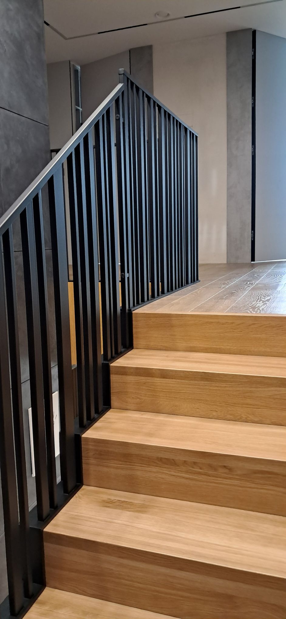 Nowoczesne schody z drewnianymi stopniami i czarną, minimalistyczną balustradą. Widok z dołu, podkreślający solidność wykonania i estetykę wykończenia.