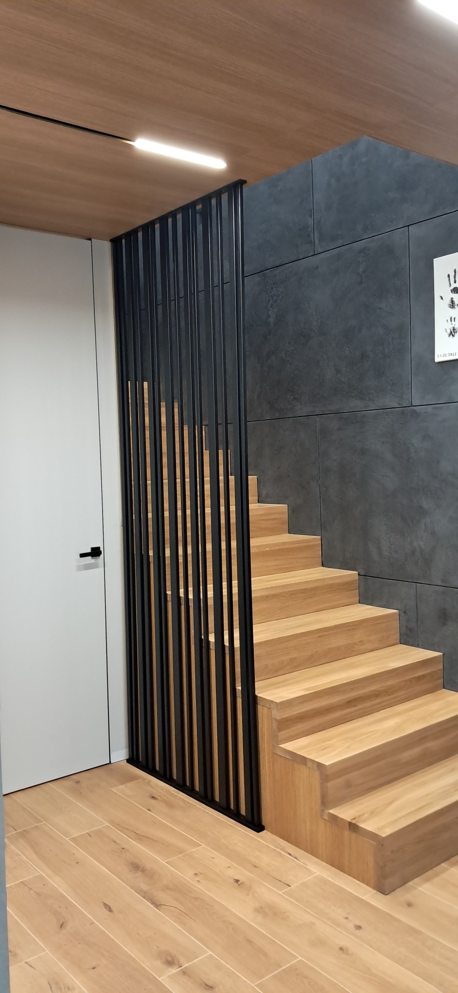 Drewniane schody z czarną, pionową balustradą w nowoczesnym wnętrzu. Jasna podłoga i ciemnoszara ściana tworzą kontrast. Minimalistyczny design.