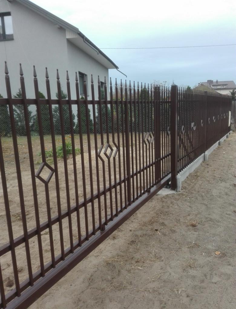 Brązowe ogrodzenie posesji z metalowymi prętami zakończonymi szpicami i romboidalnymi dekoracjami, widoczne na tle domu jednorodzinnego.