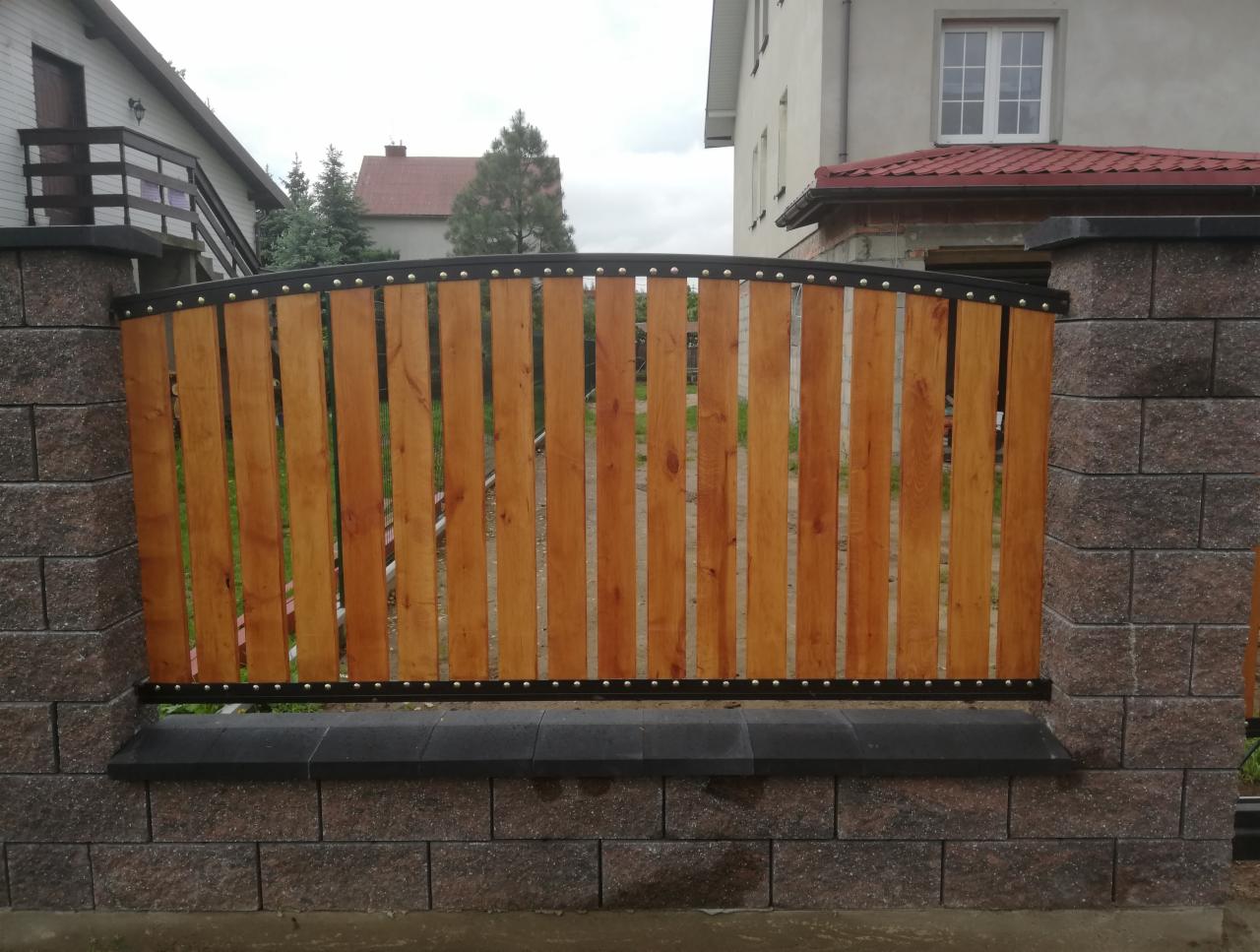 Fragment ogrodzenia z drewnianymi, pionowymi panelami w metalowej ramie, osadzonego między kamiennymi słupkami. Widoczne elementy mocujące i tło z budynkami mieszkalnymi.