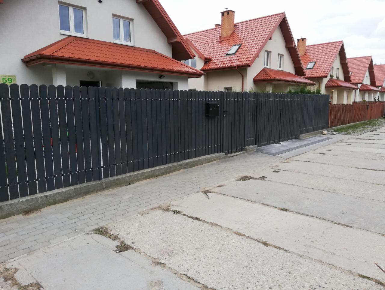 Ciemnoszare ogrodzenie panelowe z furtką i skrzynką na listy, widok na szereg domów z czerwonymi dachami