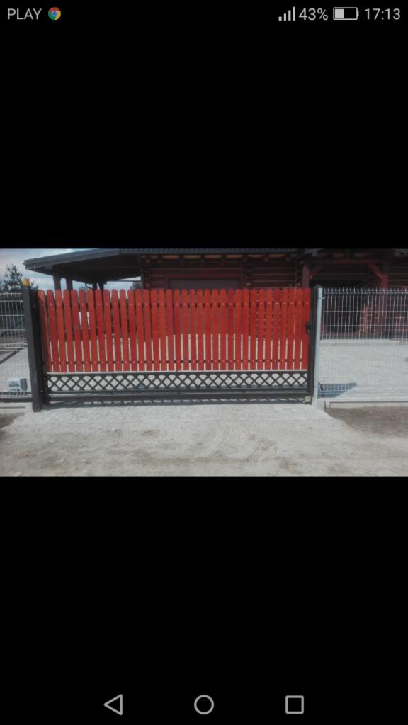 Czerwona drewniana brama wjazdowa z ozdobnym czarnym ażurem u dołu, obok panel ogrodzeniowy z siatki.