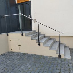 Damian Kanik Wykończenia wnętrz - Zewnętrzne schody z szarego kamienia z metalową balustradą, prowadzące do drzwi wejściowych budynku, z widoczną klapą rewizyjną w podstawie schodów.