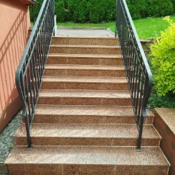 Zewnętrzne schody z brązowego granitu z czarną, kutą balustradą, prowadzące do wejścia budynku, otoczone zielenią.