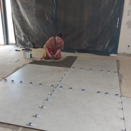 Damian Kanik Wykończenia wnętrz - Układanie wielkoformatowych płytek podłogowych z użyciem krzyżyków dystansowych; glazurnik w trakcie pracy, widoczne narzędzia i materiały budowlane.