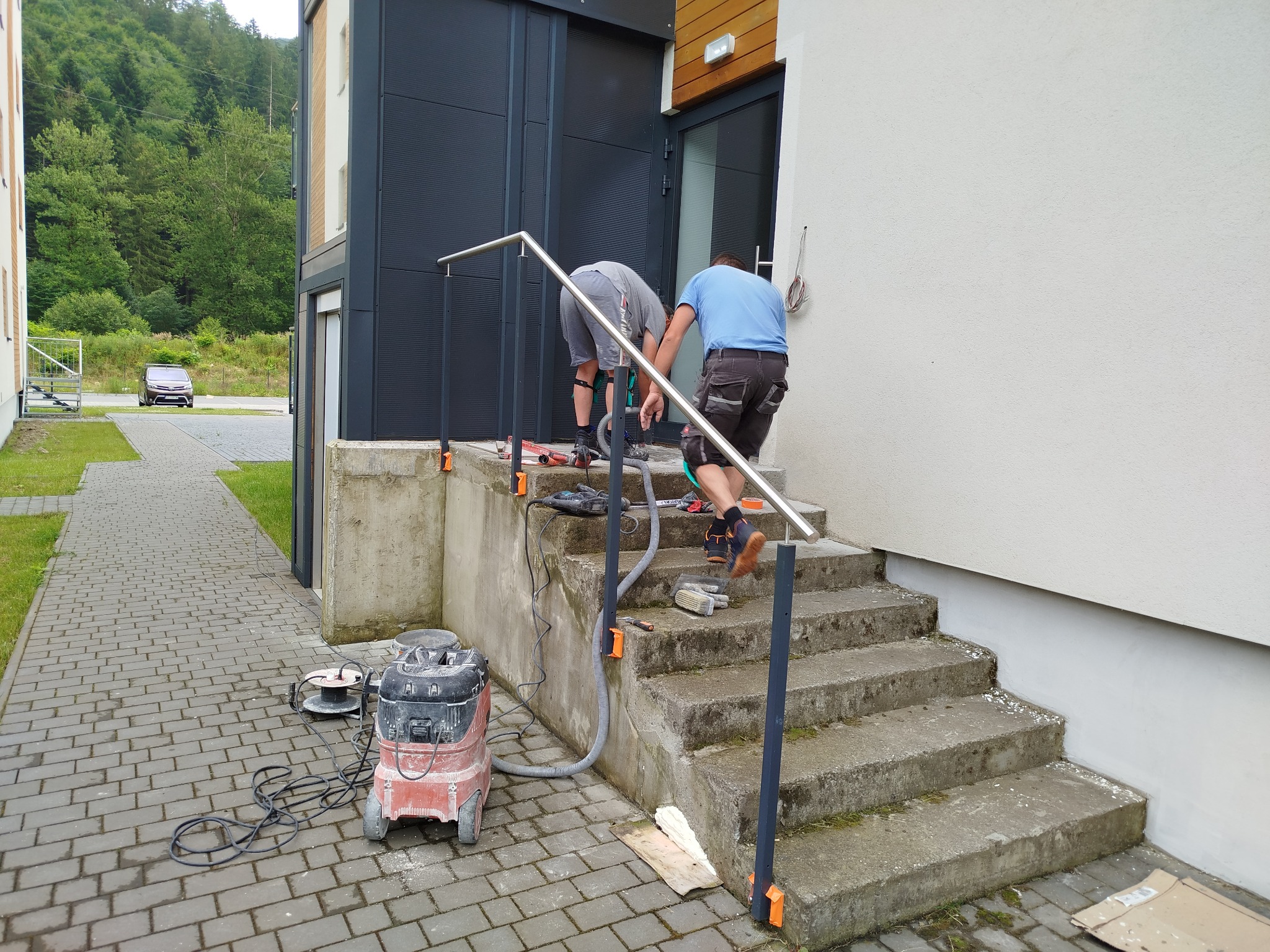 Dwóch pracowników montuje metalową barierkę na betonowych schodach zewnętrznych prowadzących do budynku. Widoczny sprzęt budowlany, w tym odkurzacz przemysłowy i narzędzia.