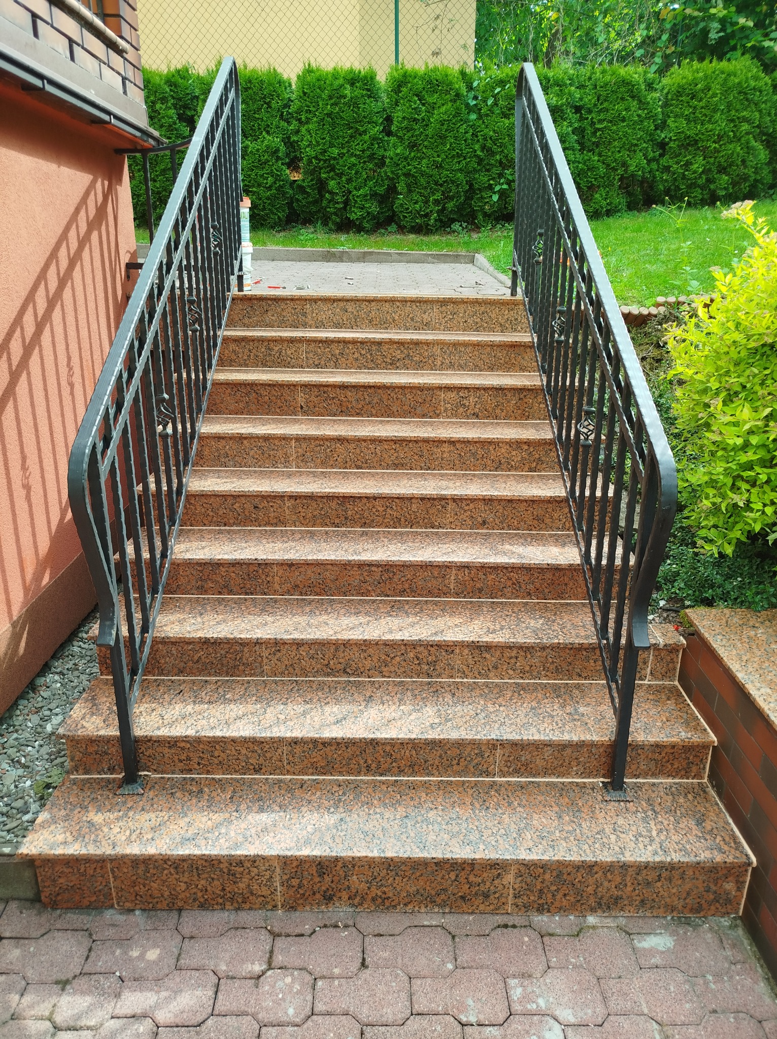 Zewnętrzne schody z brązowego granitu z czarną, kutą balustradą, prowadzące do wejścia budynku, otoczone zielenią.