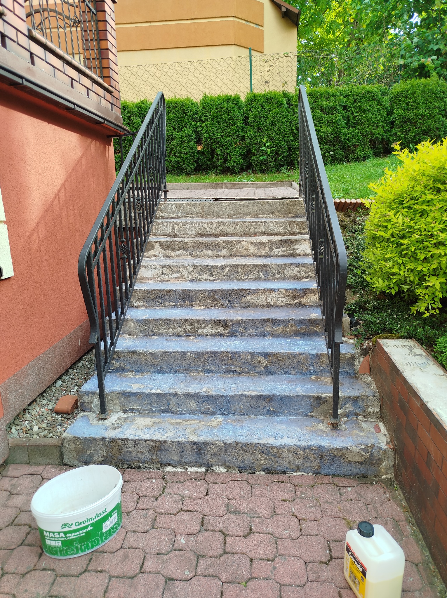 Betonowe schody zewnętrzne z czarną metalową balustradą, widoczne ślady starej farby, obok wiadro i butelka z preparatem budowlanym.