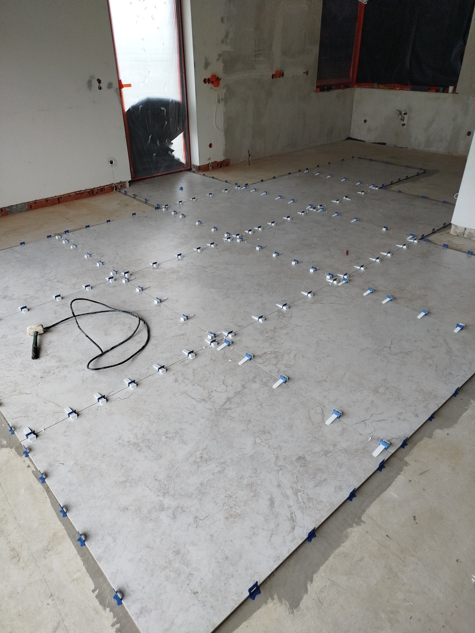 Układanie szarych płytek podłogowych z użyciem systemu poziomowania. Widoczne narzędzia glazurnicze i zabezpieczone folią drzwi w surowym wnętrzu.