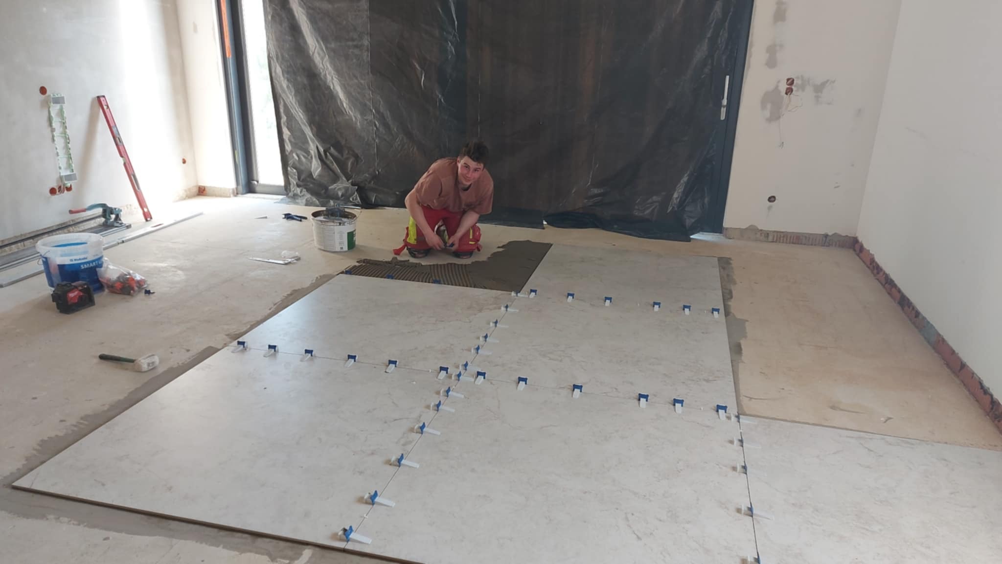 Układanie wielkoformatowych płytek podłogowych z użyciem krzyżyków dystansowych; glazurnik w trakcie pracy, widoczne narzędzia i materiały budowlane.