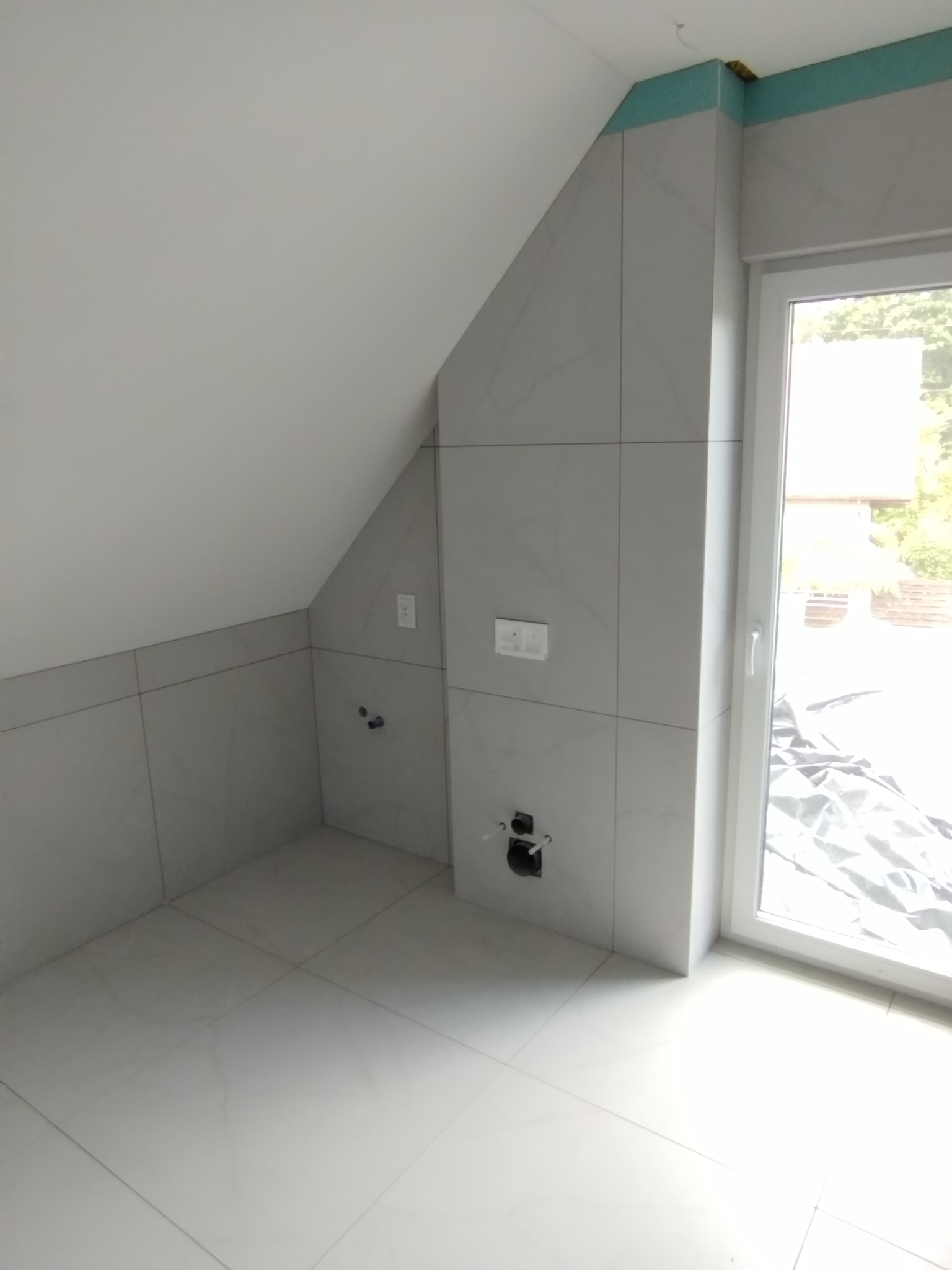 Wykończona łazienka na poddaszu ze skośnym sufitem, wyłożona jasnymi, marmurkowymi płytkami. Widoczne przyłącza hydrauliczne i elektryczne. Okno z widokiem na zewnątrz.