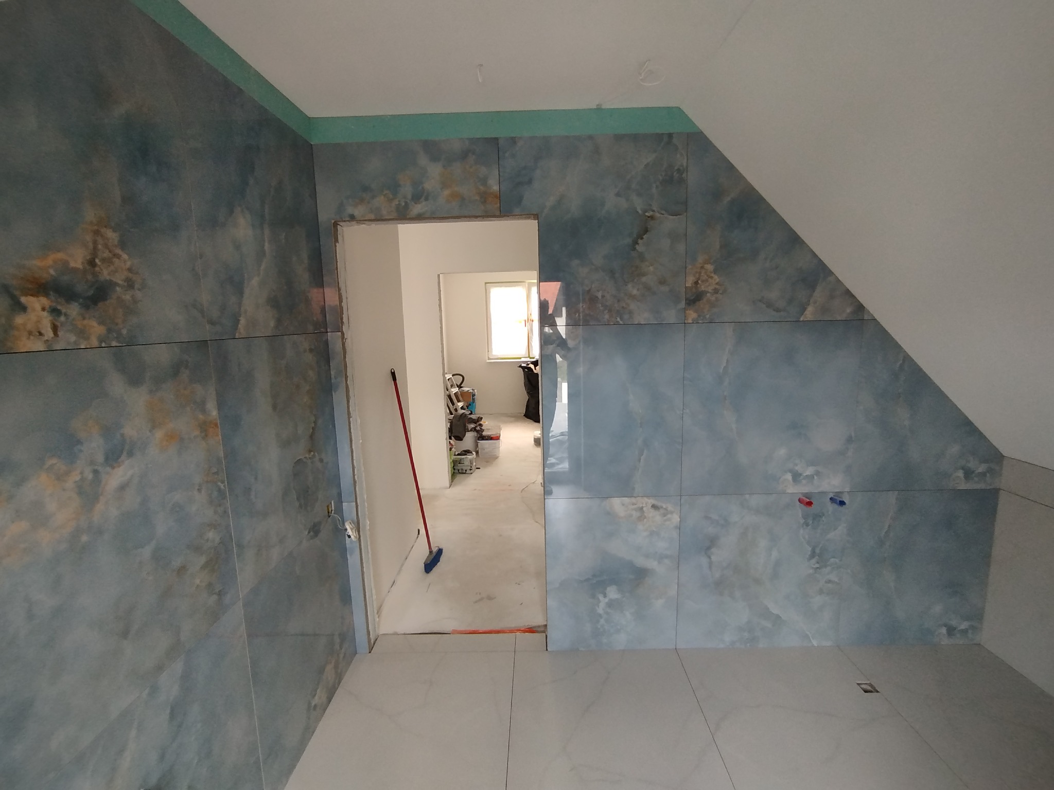 Niebieskie płytki imitujące marmur ułożone na ścianach i podłodze w łazience na poddaszu. Widoczny fragment ściany z oknem w głębi pomieszczenia.