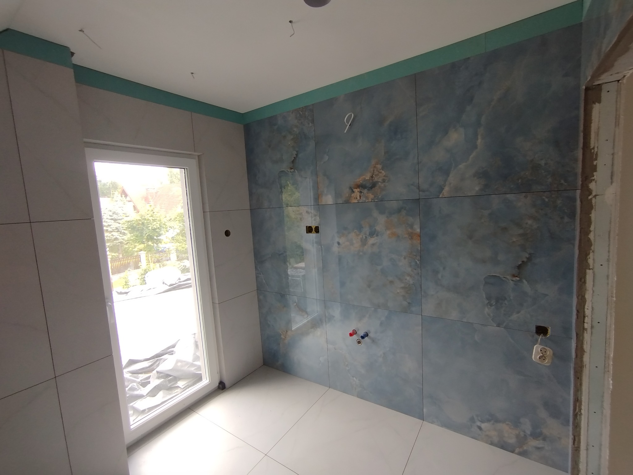 Wykończona łazienka z płytkami imitującymi marmur, widoczne przyłącza wodne i gniazdko elektryczne. Okno z widokiem na zewnątrz. Całość utrzymana w jasnej tonacji.