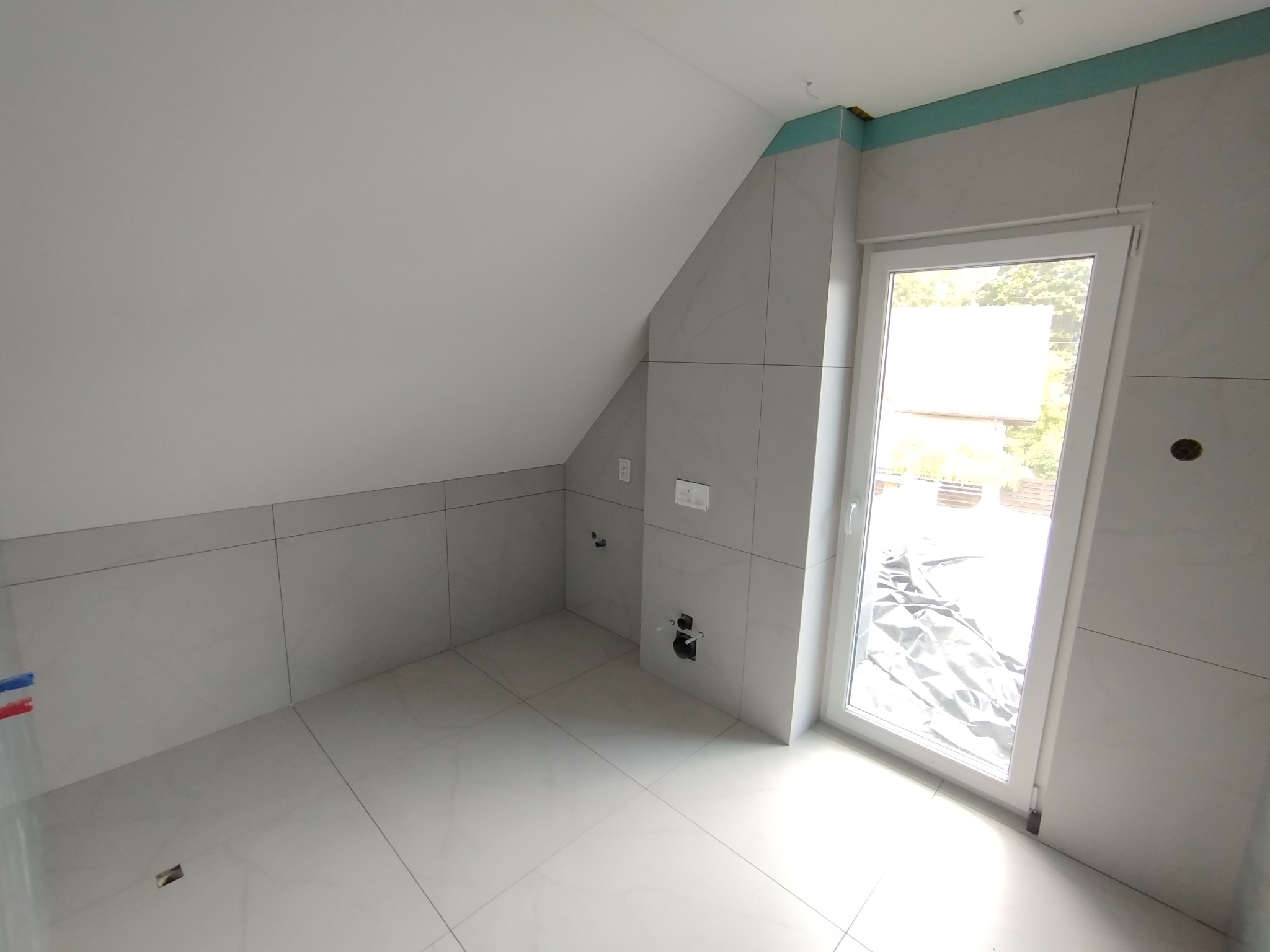 Wykończone wnętrze poddasza z jasnymi, marmurkowymi płytkami na podłodze i ścianach. Widoczne instalacje, okno balkonowe i skosy sufitu.