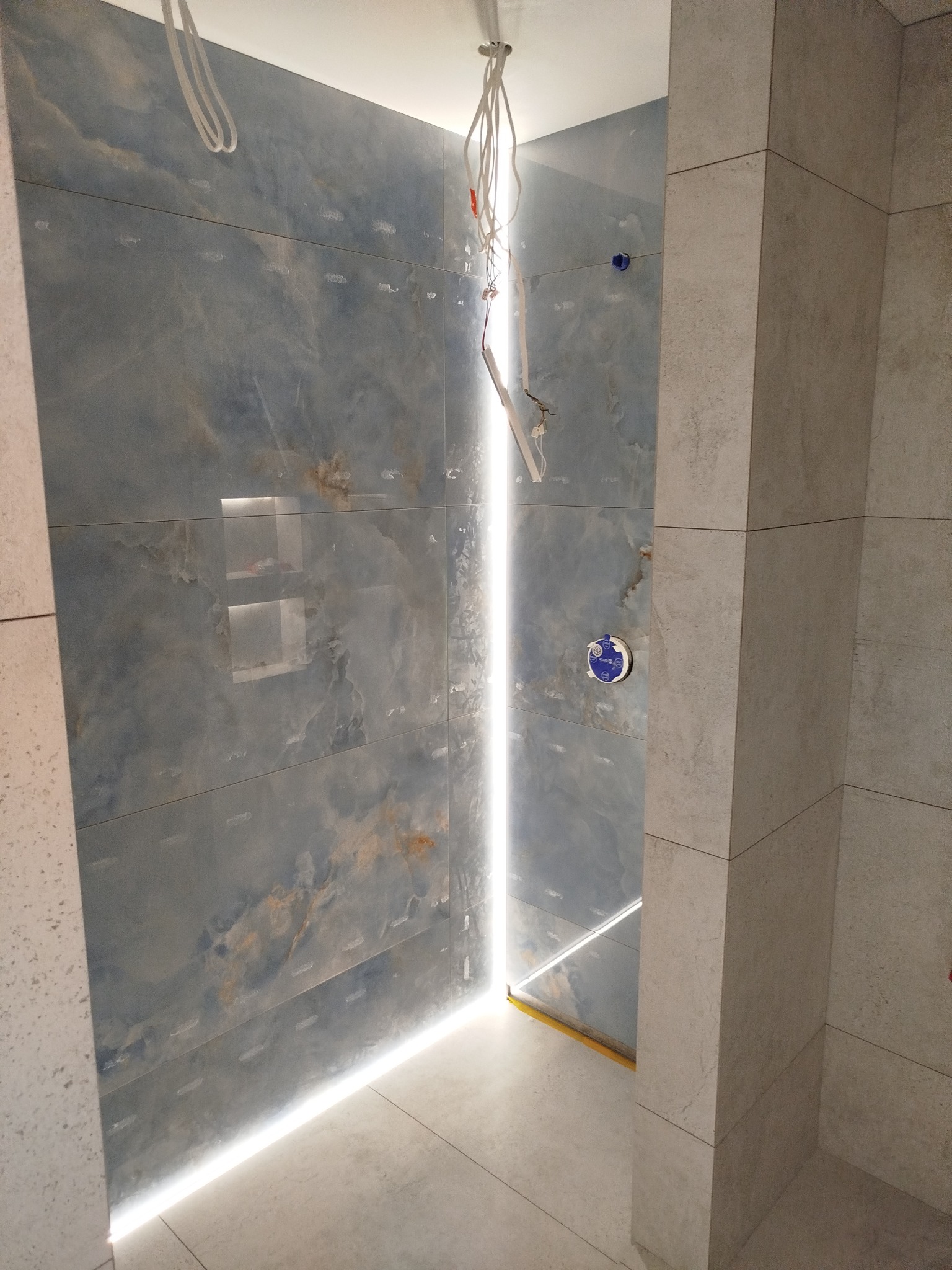 Wykończona łazienka z niebieskimi płytkami imitującymi marmur i subtelnym oświetleniem LED wzdłuż podłogi. Widoczne kable i instalacje przed ostatecznym wykończeniem.