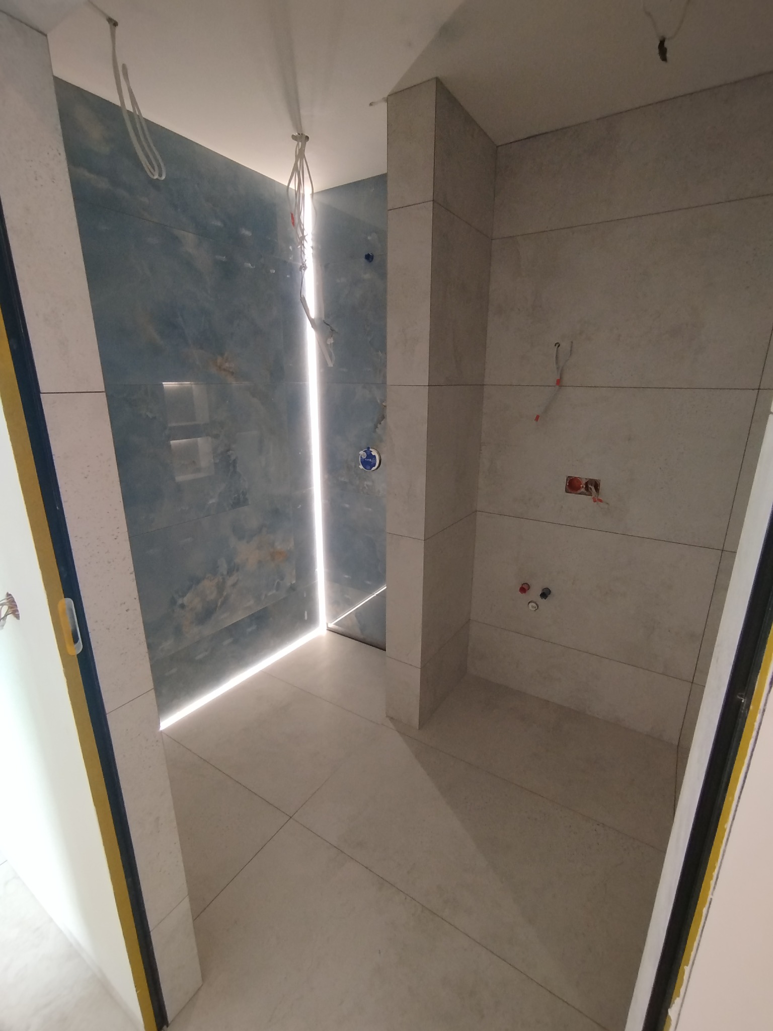 Wykończona łazienka z niebieskimi i szarymi płytkami. Widoczne instalacje wodne i elektryczne. Podświetlenie LED w strefie prysznica.