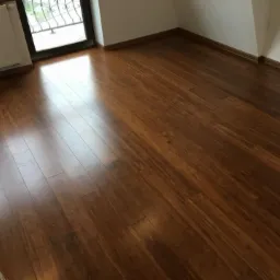 Świeżo ułożona, lśniąca, brązowa podłoga z paneli w pustym pokoju, widoczne wykończeniowe listwy przypodłogowe i fragment grzejnika pod oknem.