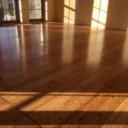 Świeżo wycyklinowana, lakierowana podłoga z desek w pustym pomieszczeniu, oświetlona promieniami słońca wpadającymi przez okno, widoczne refleksy światła.