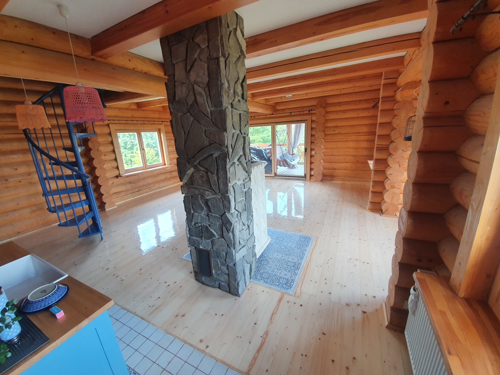 Wnętrze domu z bali z drewnianą podłogą i kamiennym kominem. Widoczna niebieska spiralna klatka schodowa i fragment kuchni z niebieskimi szafkami.