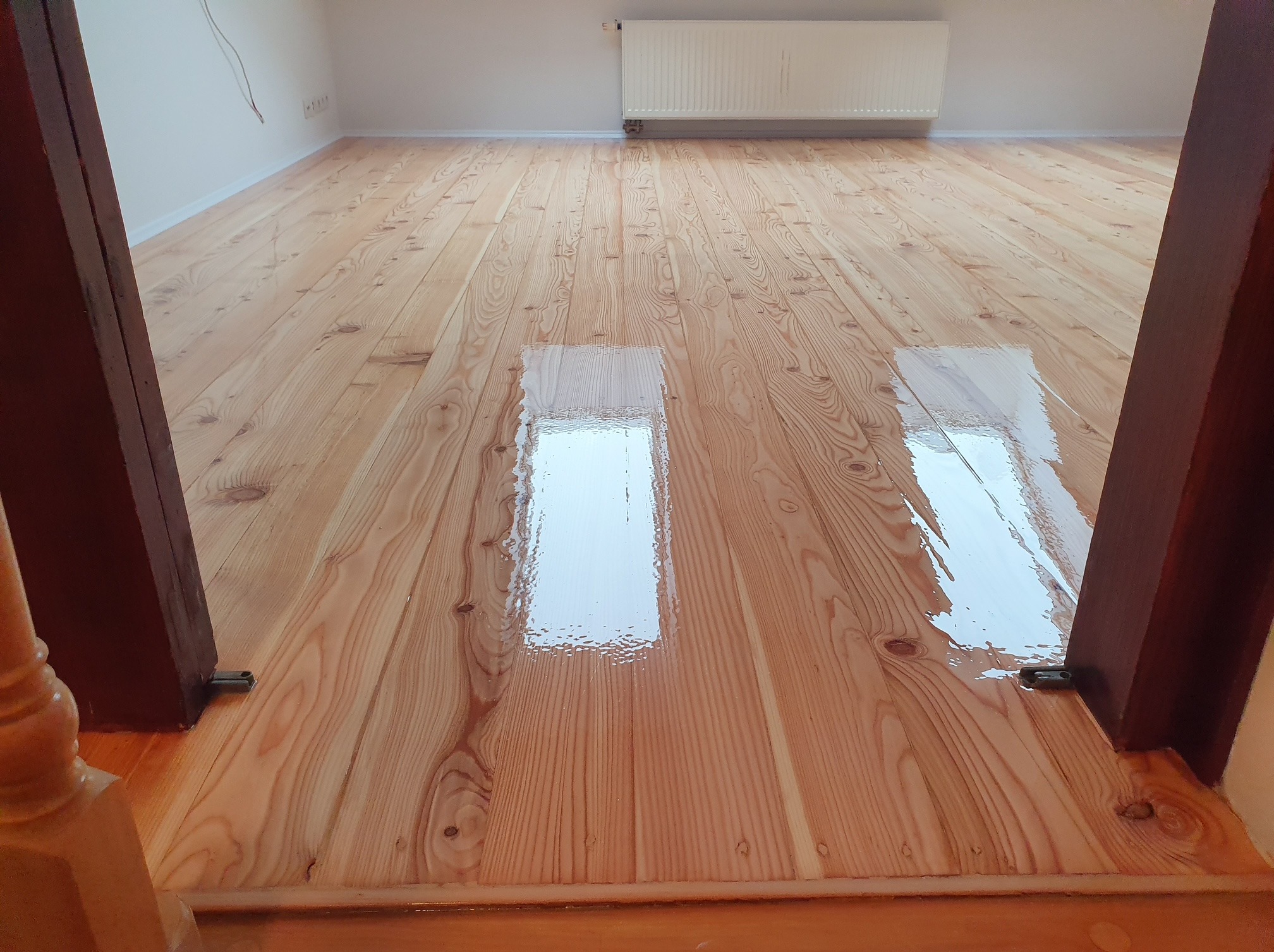 Błyszcząca, świeżo polakierowana podłoga z jasnego drewna, widziana z perspektywy progu. Widoczne odbicia światła na gładkiej powierzchni i fragment grzejnika w tle.