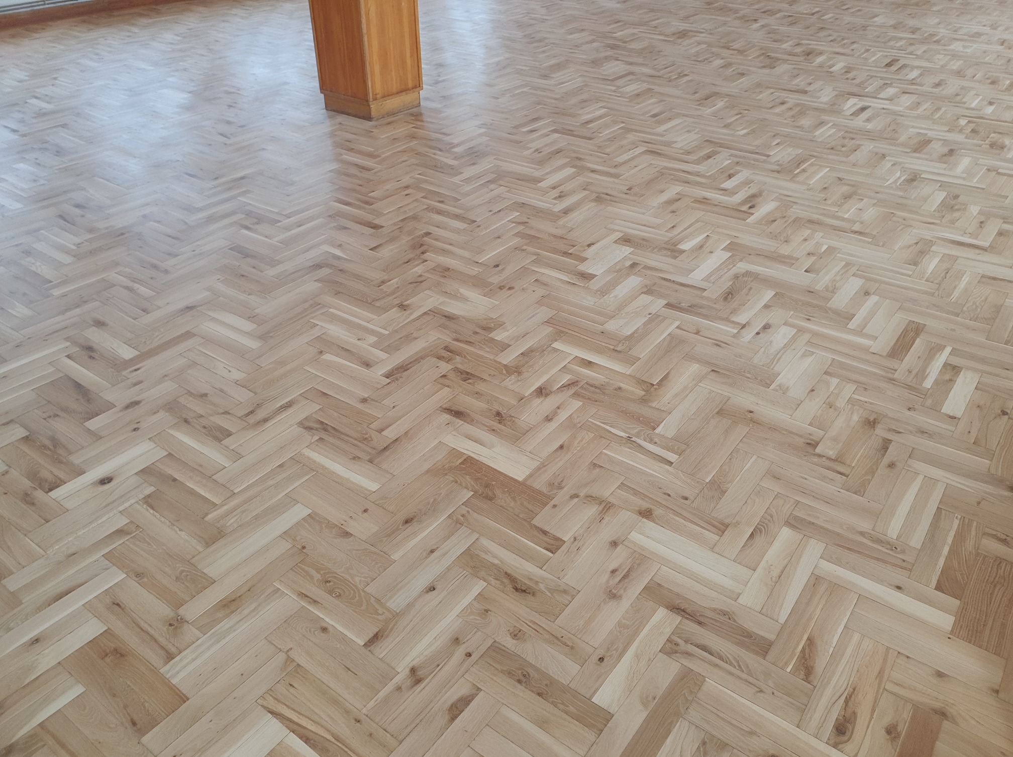 Jasny parkiet ułożony we wzór jodełki francuskiej, z widoczną kolumną w rogu. Błyszcząca powierzchnia odbija światło, podkreślając naturalne usłojenie drewna.