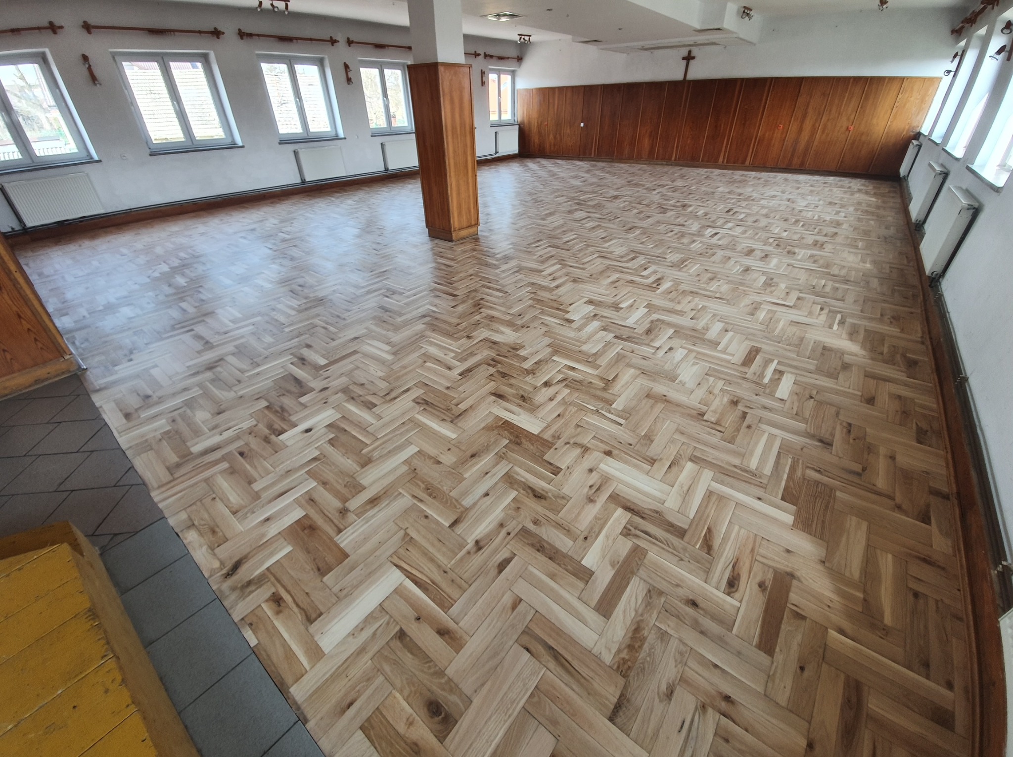 Nowa, jasna podłoga z parkietu ułożonego we wzór jodełki w przestronnym, jasnym pomieszczeniu z oknami i drewnianą boazerią. Widoczny fragment schodów.