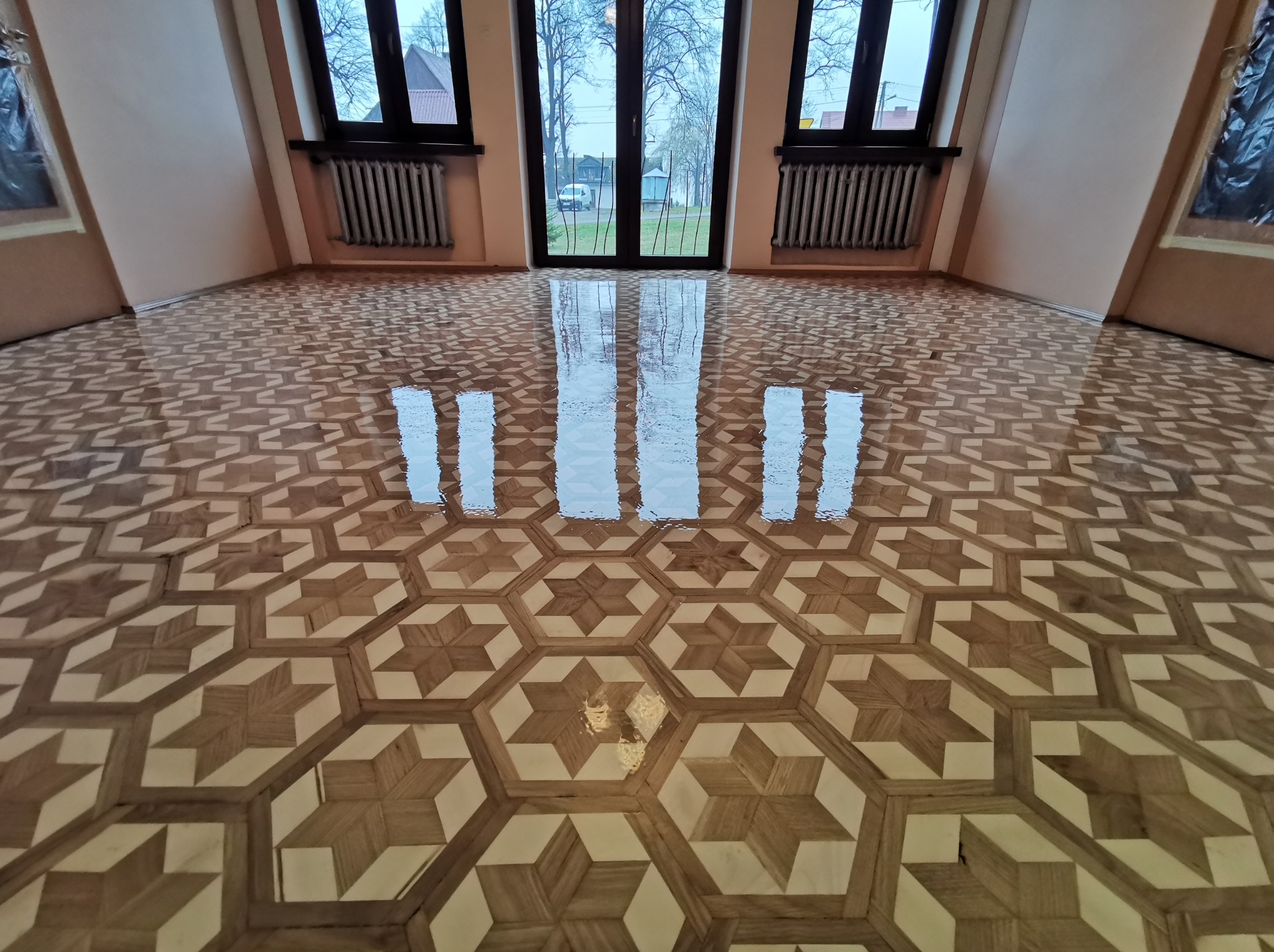 Wzorzysta, błyszcząca podłoga parkietowa w jodełkę, świeżo po renowacji w jasnym pomieszczeniu w Gdowie. Widoczne odbicia w lakierze i okna.