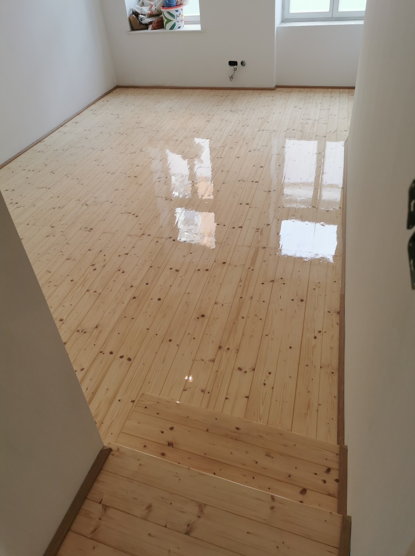 Jasne, lakierowane deski podłogowe z widocznymi słojami drewna, schody wykończone tym samym materiałem, białe ściany, okno w tle. Widok z góry, świeżo po lakierowaniu.