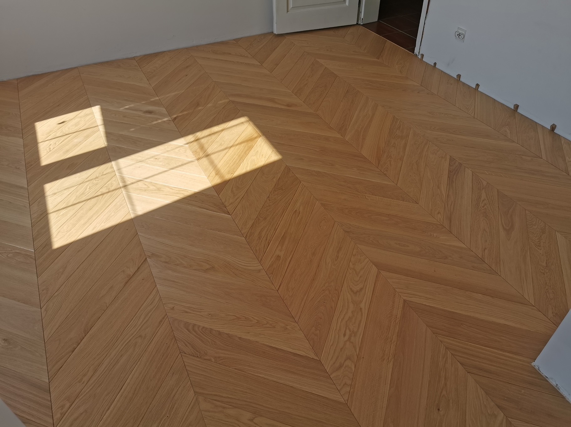 Jasny, drewniany parkiet ułożony we wzór jodełki francuskiej, oświetlony promieniami słońca wpadającymi przez okno. Widoczne kołki dystansowe przy ścianie.