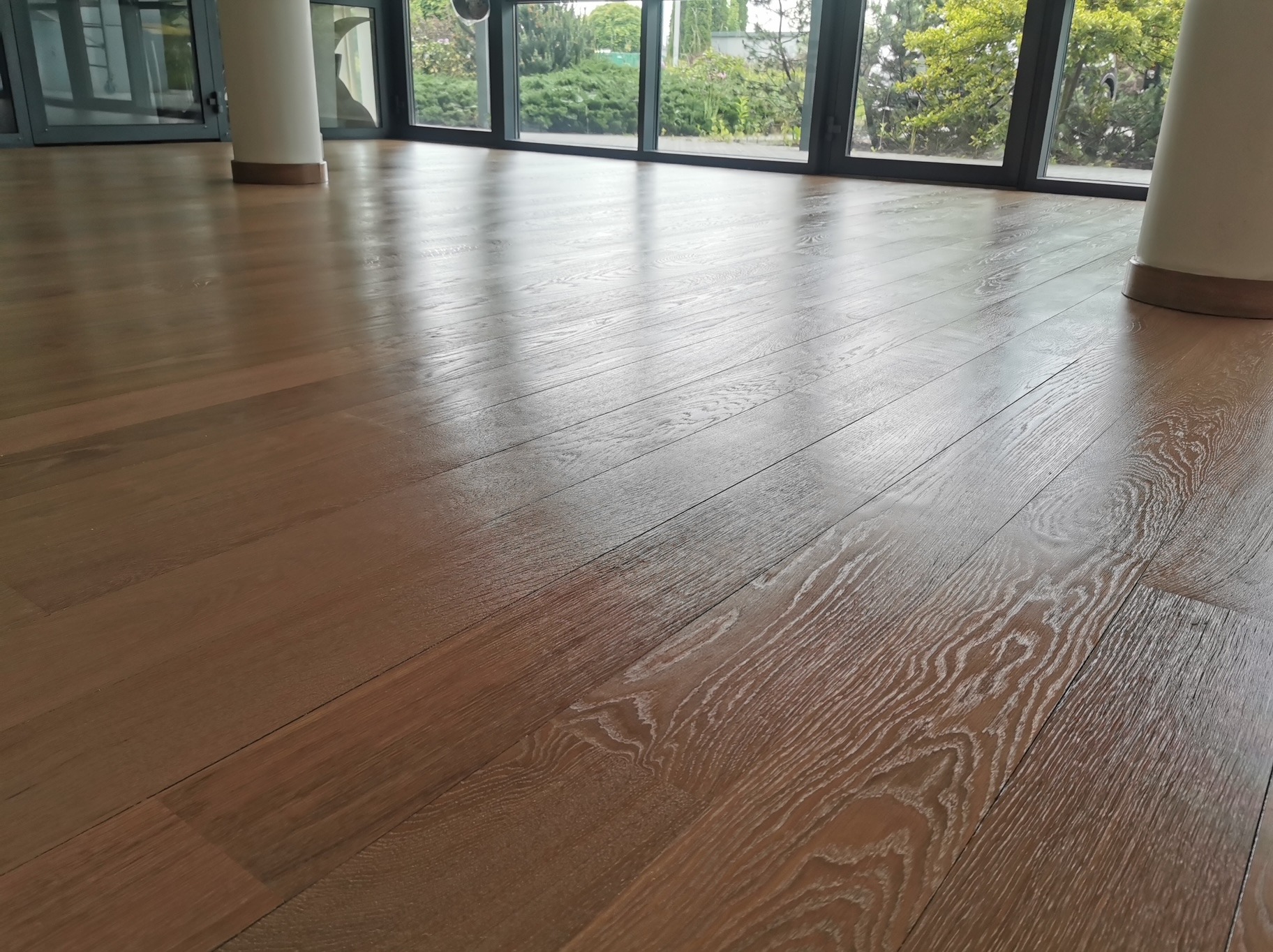 Położony parkiet w jasnym odcieniu brązu, z widoczną strukturą drewna, w przestronnym wnętrzu z dużymi oknami i kolumnami. Widok na zieleń za oknem.