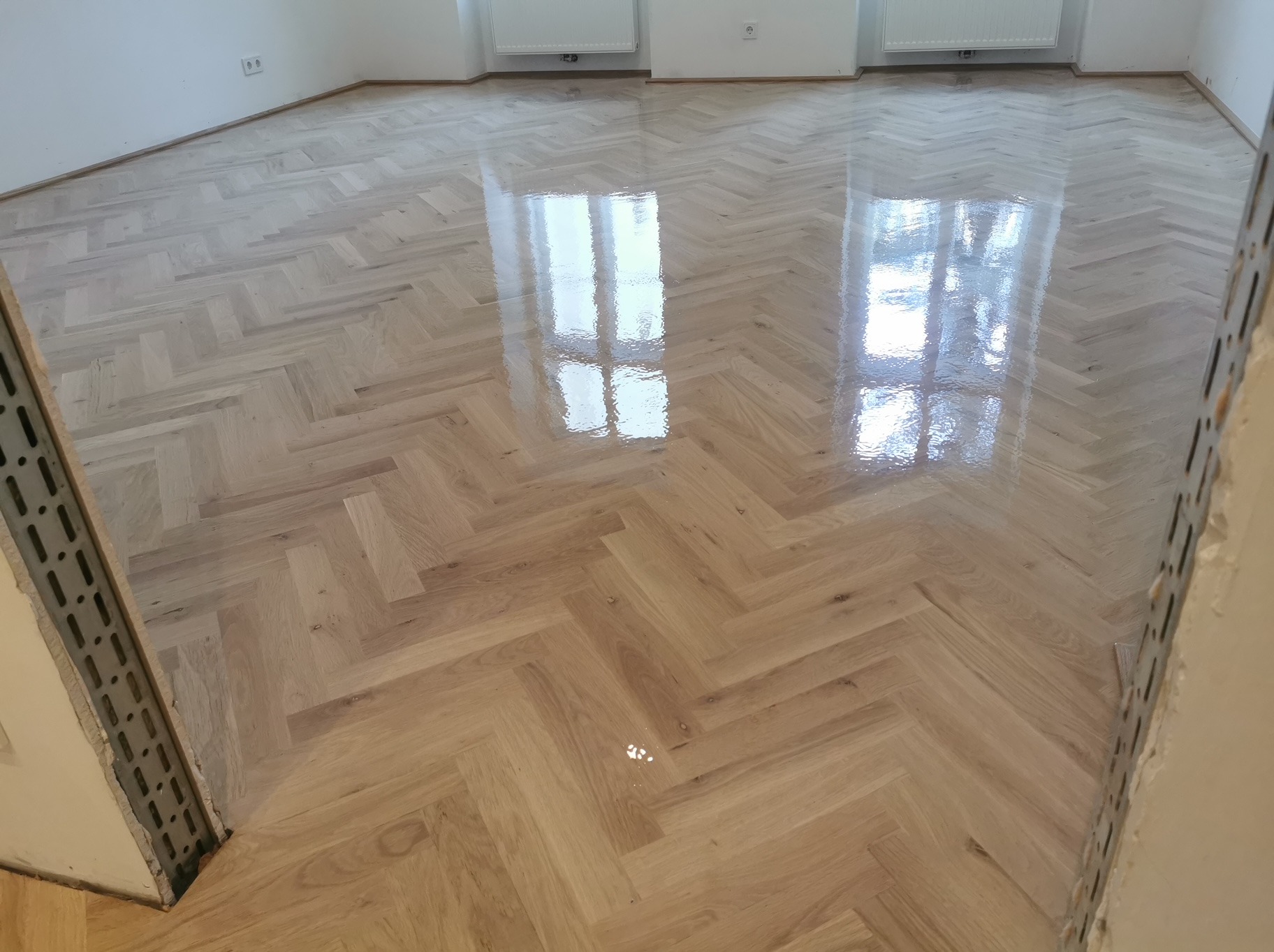 Błyszcząca, jasna podłoga z parkietu ułożonego w jodełkę, odbijająca światło okien, widoczna z perspektywy narożnika remontowanego pomieszczenia.