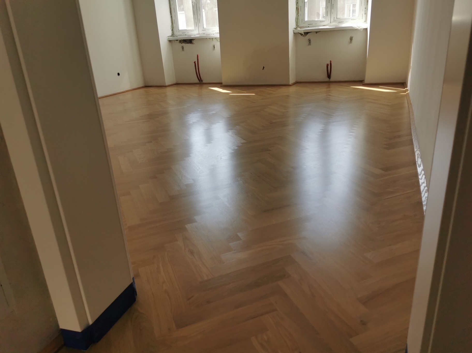 Jasny parkiet ułożony w jodełkę w pustym pokoju z oknami, widok zza framugi drzwi. Podłoga po renowacji, z widocznym połyskiem.