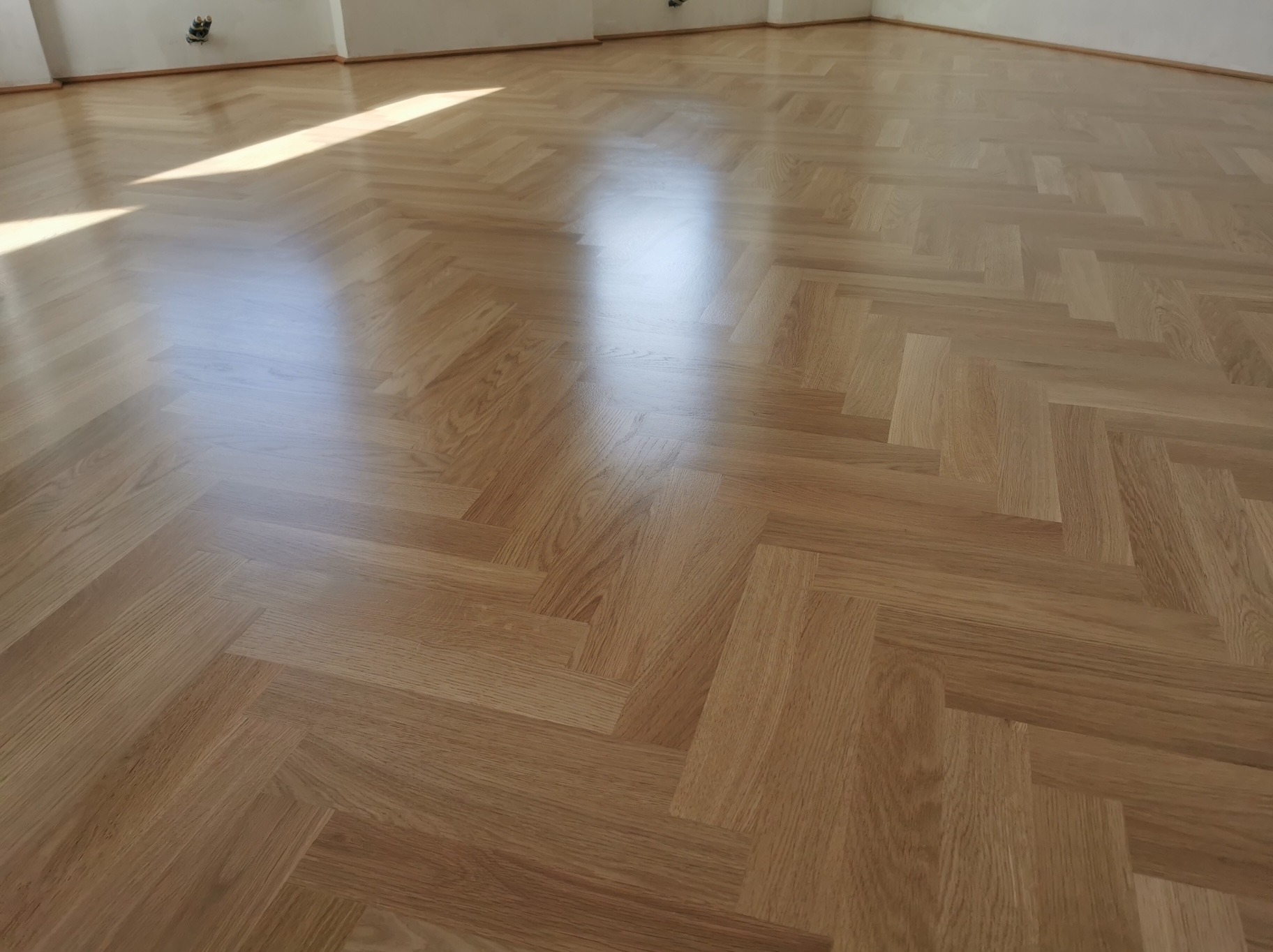 Parkiet ułożony w jodełkę, świeżo po cyklinowaniu, z widocznym refleksem światła słonecznego. Jasne drewno, gładka powierzchnia, minimalistyczne wnętrze.