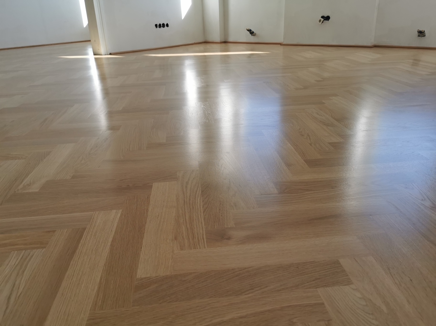 Błyszcząca, jasna podłoga z parkietu ułożonego we wzór jodełki francuskiej. Świeże wykończenie widoczne w odbiciu światła. Białe ściany i widoczne gniazdka elektryczne.